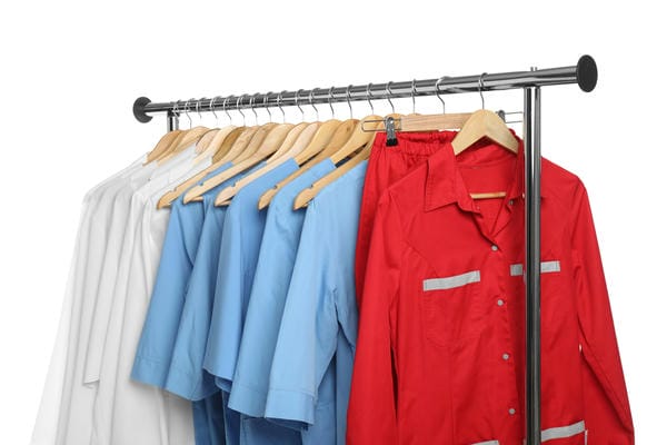 arbeitsbedingungen-berufskleidung Eine Auswahl an Berufsbekleidung, bestehend aus weißen, hellblauen und roten Hemden sowie einer roten Hose, hängt ordentlich auf Holzbügeln an einer metallischen Kleiderstange.