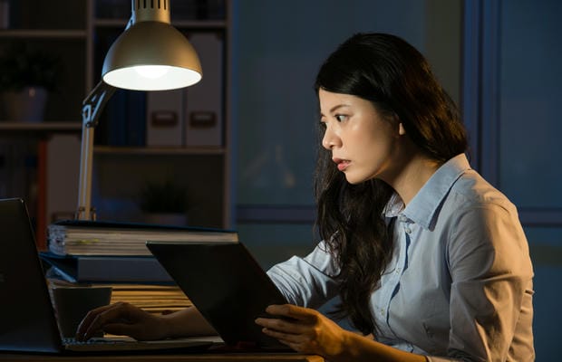 arbeitsbedingungen-ueberstunden Eine konzentrierte Frau macht nachts unter einer Schreibtischlampe Überstunden, während sie gleichzeitig an einem Laptop und einem Tablet arbeitet.