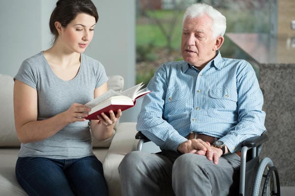 Chancen und Risiken als Sozialarbeiter junge Frau liest altem Mann im Rollstuhl ein Buch vor.