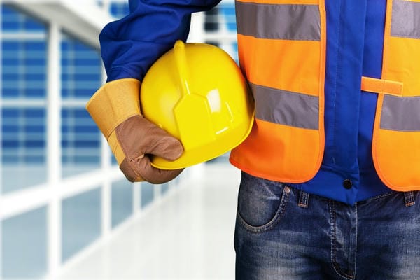 arbeitsplatz-arbeitsschutz Ein Bauarbeiter in blauer Arbeitskleidung und orangefarbener Warnweste hält als Zeichen für den Arbeitsschutz einen gelben Schutzhelm unter dem Arm.