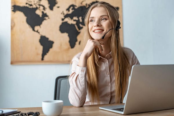 arbeitsbedingungen-vertrag-rechte-pflichten Eine Tourismuskauffrau mit Headset vorm Laptop. Im Hintergrund ist eine Weltkarte zu sehen.