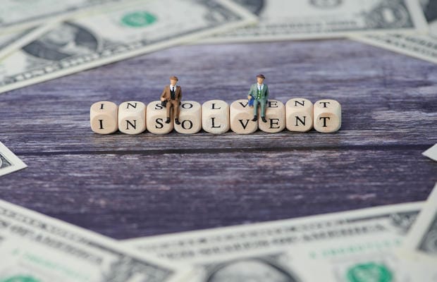insolvenz-insolvenzgeld Zwei Miniatur-Geschäftsleute sitzen deprimiert auf Holzklötzchen, die das Wort „Insolvent“ bilden und von Dollar-Geldscheinen umgeben sind, was die Notwendigkeit von Insolvenzgeld für Arbeitnehmer verdeutlicht.