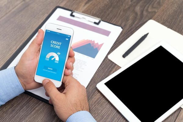 unternehmen-bonitaet-fuer-unternehmen Ein Mann überprüft seine Bonität und Kredit-Score auf einer Smartphone-App über einem Schreibtisch mit Dokumenten und einem Tablet.