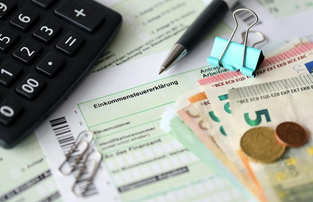 verguetung-netto-einkommensteuer Ein deutsches Formular zur Einkommensteuererklärung liegt bereit, umgeben von einem Taschenrechner, Euro-Geldscheinen und Münzen sowie einem Stift für die Berechnung des Netto-Verdienstes.